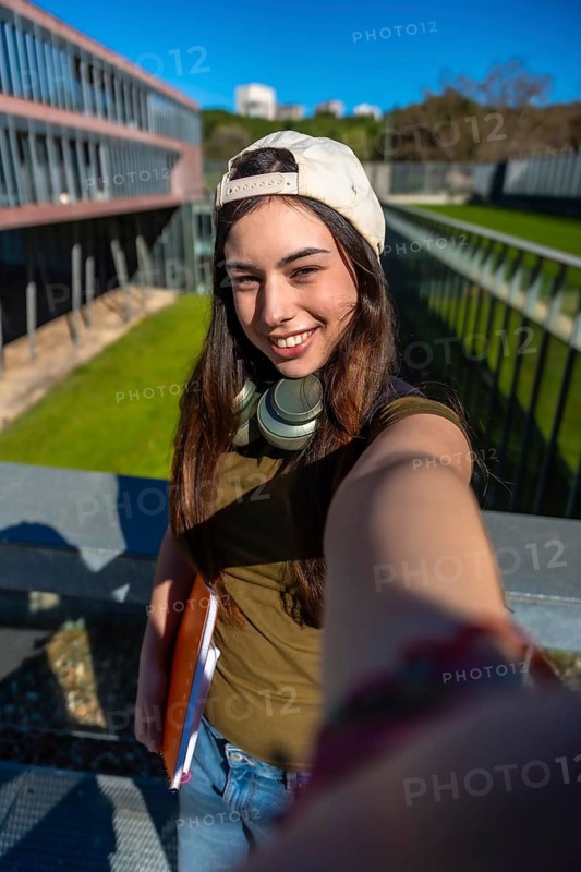 Young female student with headphones and notebooks posing for a selfie on university campus, embracing youth, education, and student lifestyle in outdoor setting during daytime