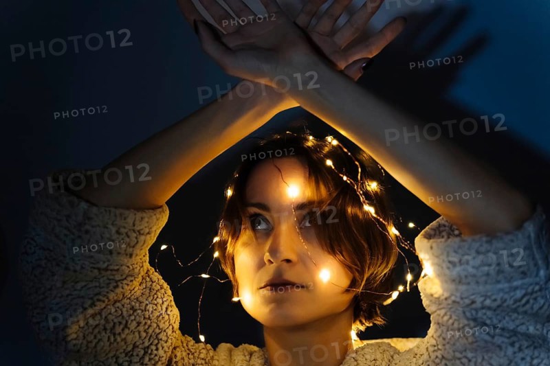 Portrait of Woman in Dark with String Lights. Woman with short hair stands in dim environment wearing textured light sweater. Warm yellow fairy lights wrapped around her head and neck, casting soft glow on her face and creating bright highlights along illuminated strands. Arms are lifted above head with crossed hands, forming graphic silhouette that projects distinct shadows onto wall behind