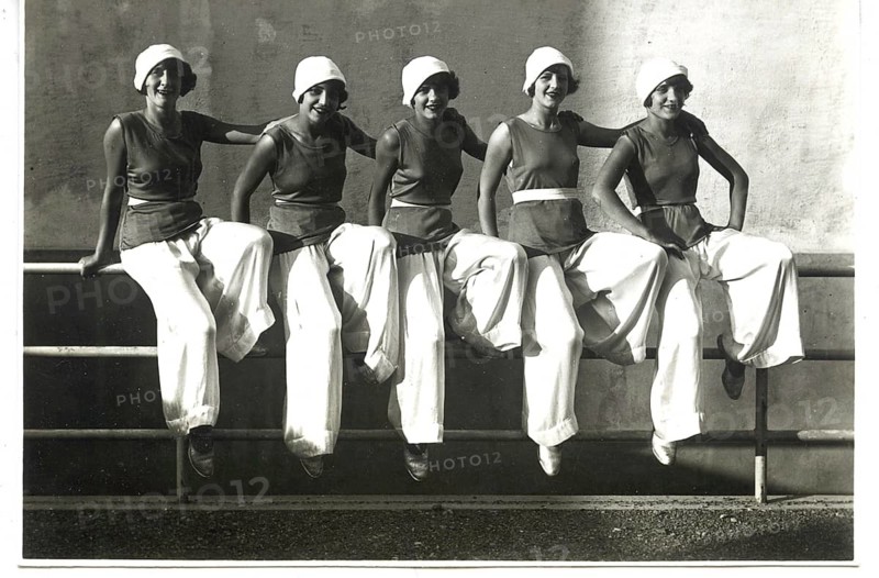 Five woman in 1920s-style clothing are sitting casually on a railing and laughing, the 1920s, self-assured, elegant white trousers (like Marlene Dietrich)