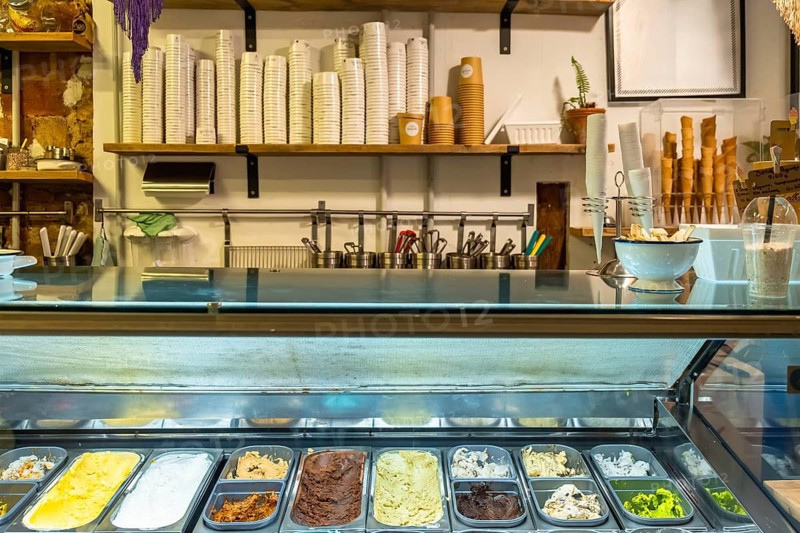 Ice cream shop interior with colorful gelato tubs in a stainless steel display case, empty cups and topping containers on shelves above the counter, ready to serve