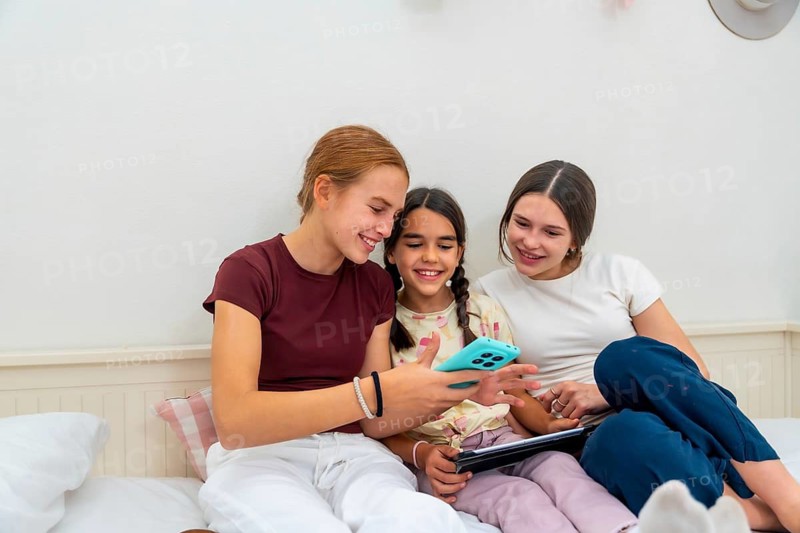 Three happy young sisters smiling and engaging with a smartphone and tablet while relaxing together on a cozy bed at home, sharing moments and connection
