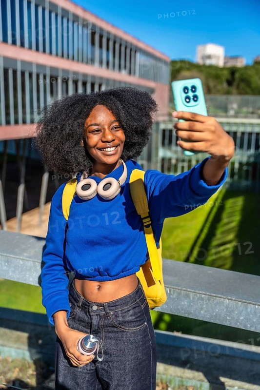 African american college student smiling on a sunny campus, taking a smartphone selfie with headphones and backpack, enjoying modern student life and social connection