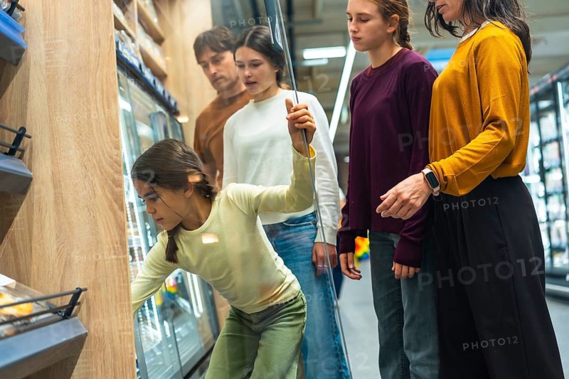 Family shopping together in a grocery store, with a young girl bending down to select items from a refrigerated display while her parents and older sister watch