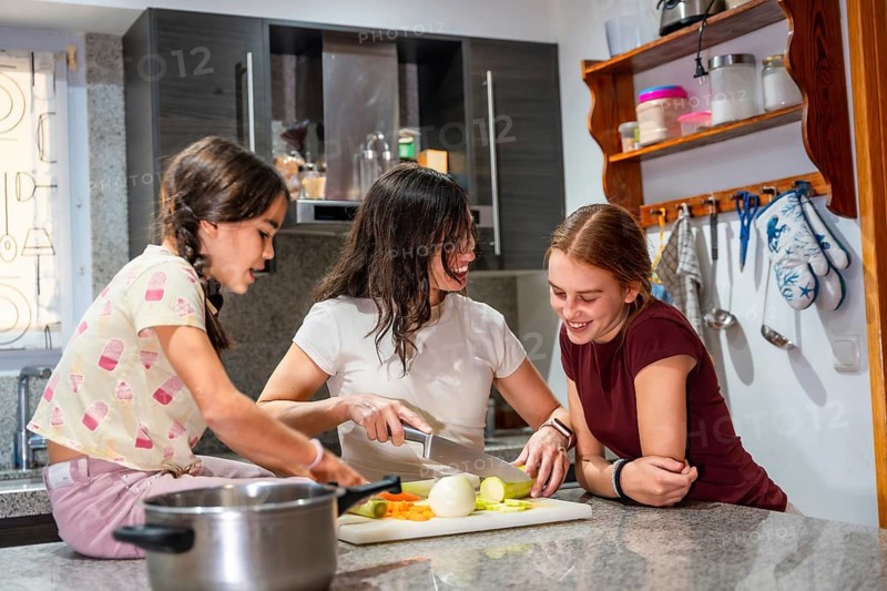 Mother and daughters smiling as they chop fresh vegetables together on a modern kitchen counter, enjoying cooking, learning, and family bonding during everyday meal prep