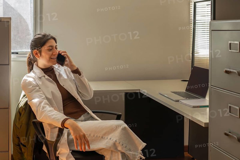 Young female scientist in white lab coat smiling while talking on a smartphone, sitting relaxed in an office chair at a desk with a laptop, filing cabinet beside her