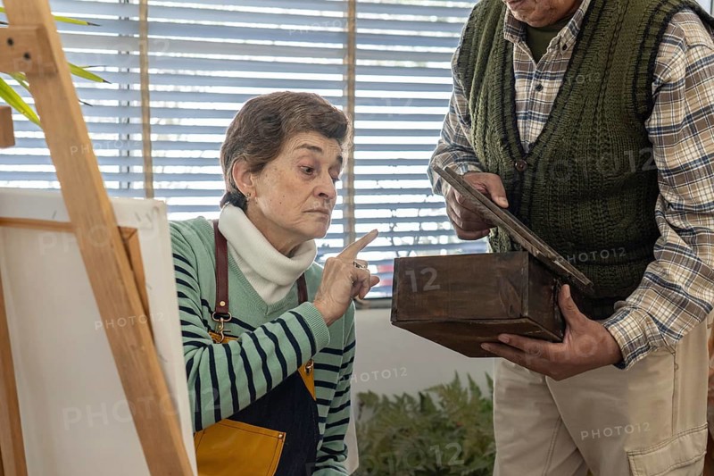 Focused older woman points at a dark wooden box held by a man, studying it carefully as reference for her artwork in a bright studio