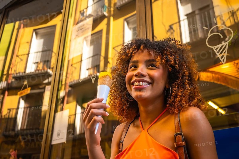 Happy young woman with curly hair enjoying an ice cream cone while smiling in an urban setting, reflecting a vibrant summer day with architectural details