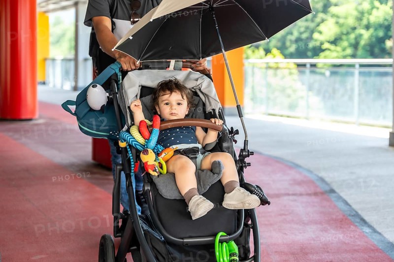 Father walks with baby son in stroller under umbrella on sunny city sidewalk, sharing protective, relaxed moments of parenting, love and exploration on a warm day