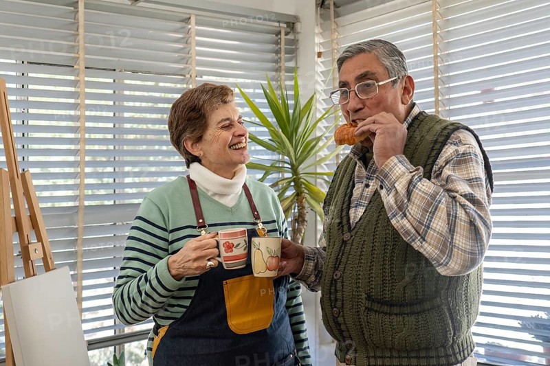 A smiling senior woman holds two colorful ceramic mugs while her husband eats a croissant beside her, both standing together and laughing in their bright home studio near a wooden easel
