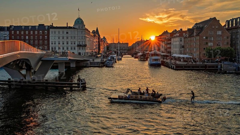 Sonnenuntergang über einer Stadt mit Brücke, Booten und einem leuchtenden goldenen Himmel, Kopenhagen, Dänemark, Nyhavn