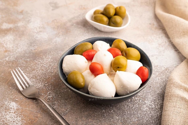 White Mozzarella cheese, with tomatoes and olives in blue bowl on brown concrete background and linen textile, side view, close up