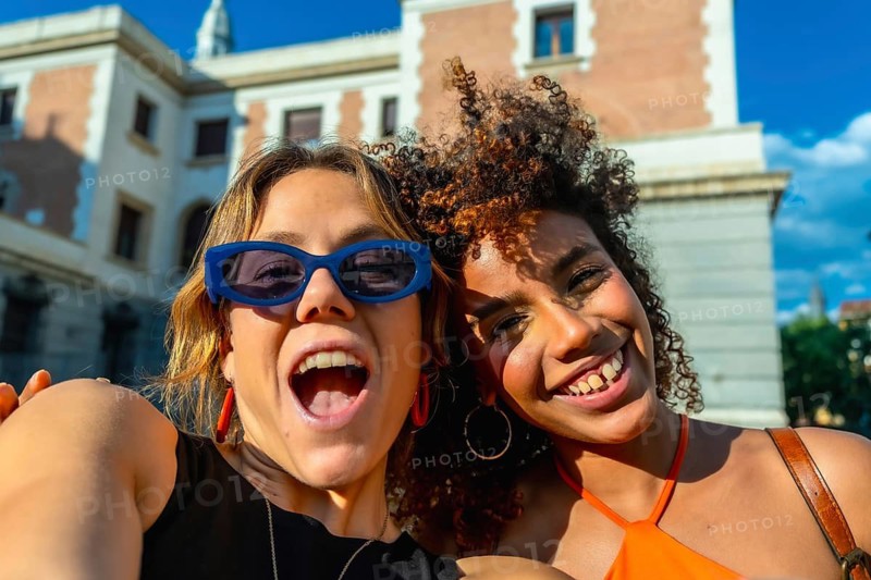 Two diverse young women smiling and laughing, capturing a joyful selfie moment in a sunny urban setting, celebrating friendship and travel experiences