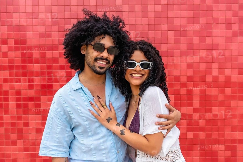 Young black couple with curly hair embracing and smiling at the camera, wearing sunglasses against a vibrant red tiled background, sharing a moment of togetherness and happiness