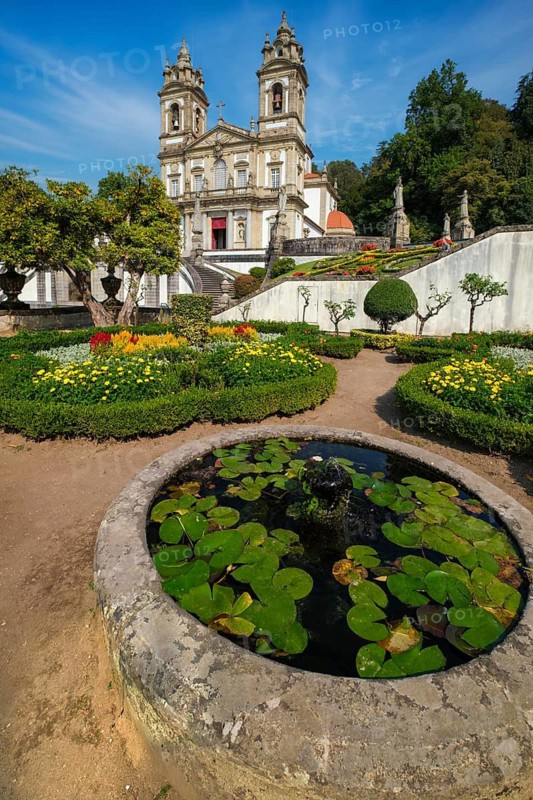 Garden view of the Bom Jesus do Monte church and sanctuary in Braga, Portugal, with a circular lily pond, manicured beds and the baroque facade rising beyond the terrace walls