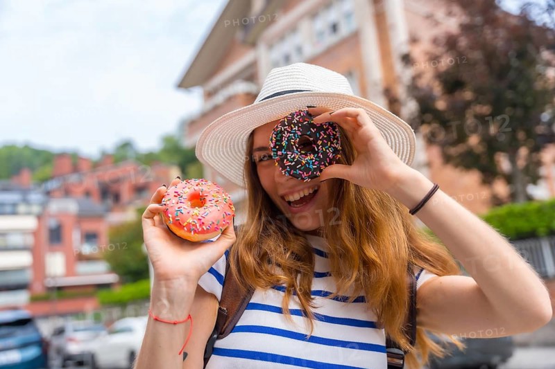 Young woman wearing a hat covering one eye with a colorful chocolate donut while holding another pink donut, smiling and enjoying a sweet snack during city travel