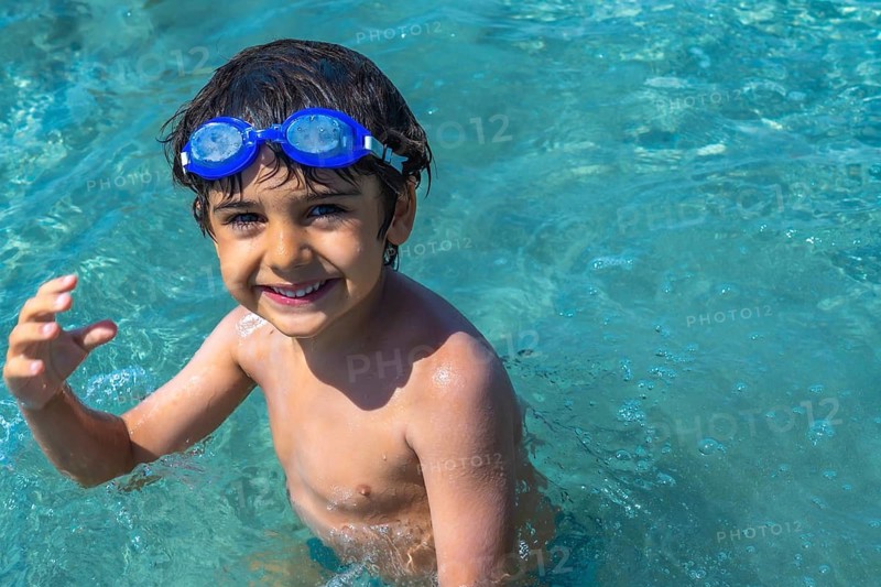 Young boy with blue swim goggles smiling at camera while swimming and playing in the clear, sparkling turquoise waters of the mediterranean sea during a refreshing summer vacation