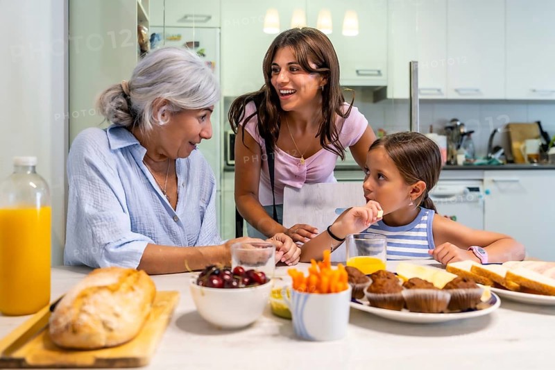 Granddaughter eating a snack, surrounded by her grandmother and mother, all three women having a cheerful family moment together in a modern kitchen