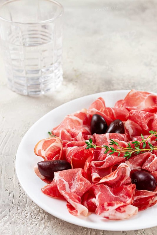 Freshly cut dried meat, served with olives and herbs, on a white plate, light background, no people