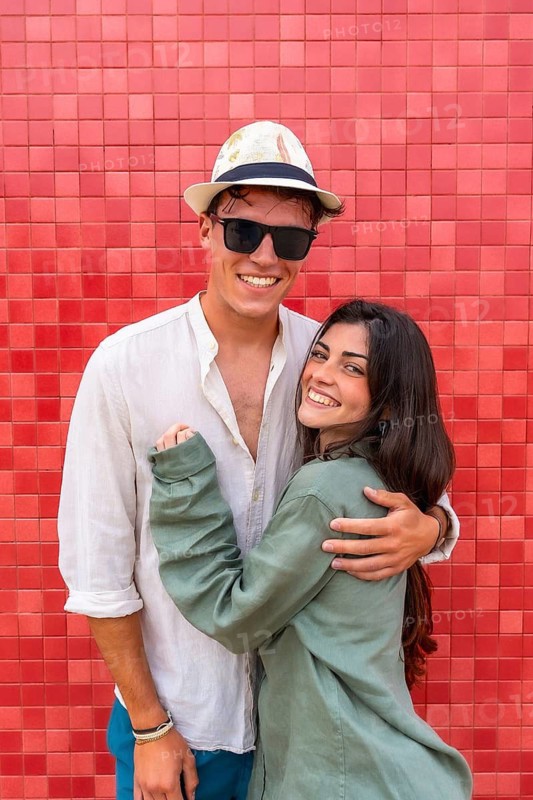 Young couple hugging and smiling against a bright red tiled wall, sharing a candid moment of affection and joy outdoors on a sunny day, relaxed and happy together
