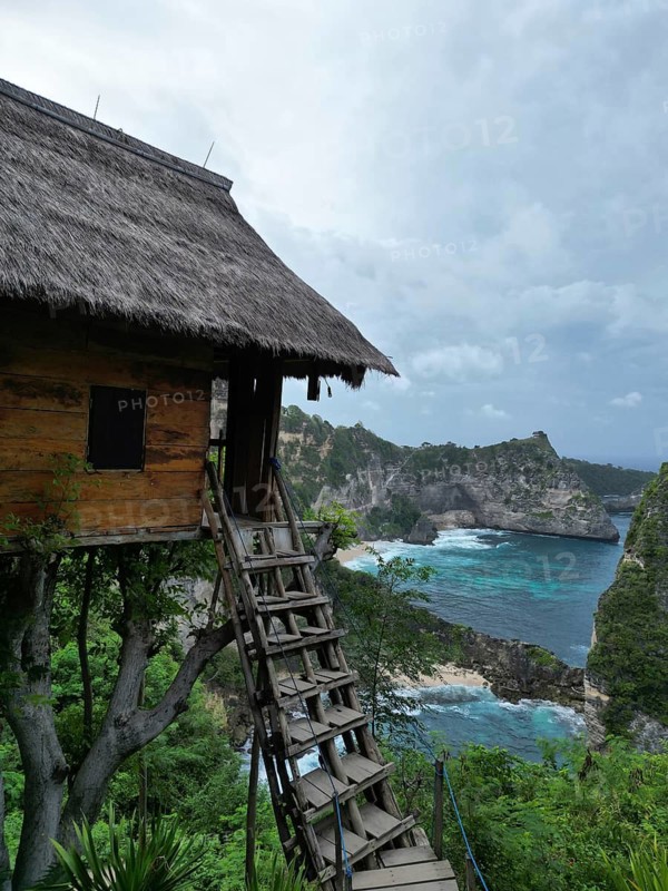 A tree house with thatched roof and wooden ladder surrounded by cliffs and a rough sea under a cloudy sky, tree house and diamond beach on the island of Nusa Penida, Bali in Indonesia