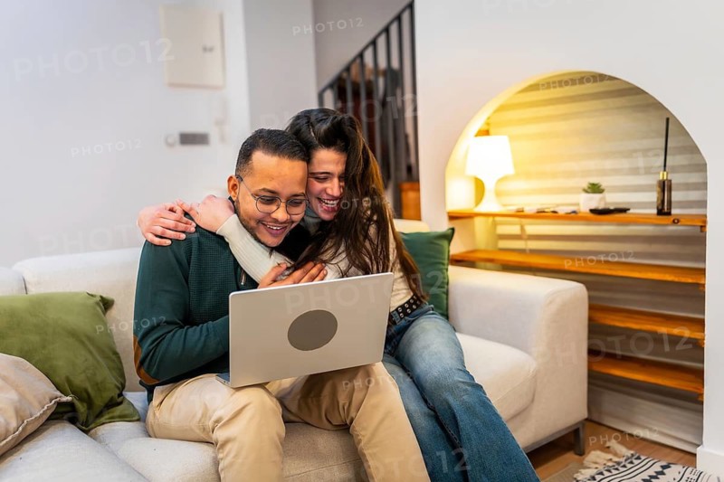 Cheerful multiethnic couple cuddling on a modern sofa, smiling and watching a movie or browsing on a laptop together in a cozy living room at home