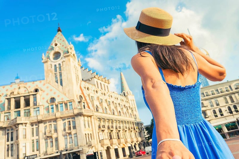 Young woman in blue dress holding partners hand walking sightseeing Europe Square in Batumi, Georgia. Neo-Gothic and Renaissance architectural style with ornate facade and spire towers