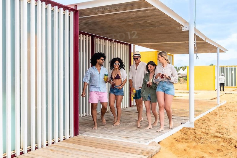 Group of diverse friends walking barefoot along a wooden pathway with refreshing drinks at a modern beach club or resort, spending sunny summer vacation time together