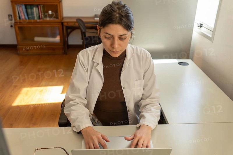 High angle view of a young female scientist in white lab coat typing on a laptop at a bright office desk, with a wooden bookcase and warm sunlight on the floor behind her