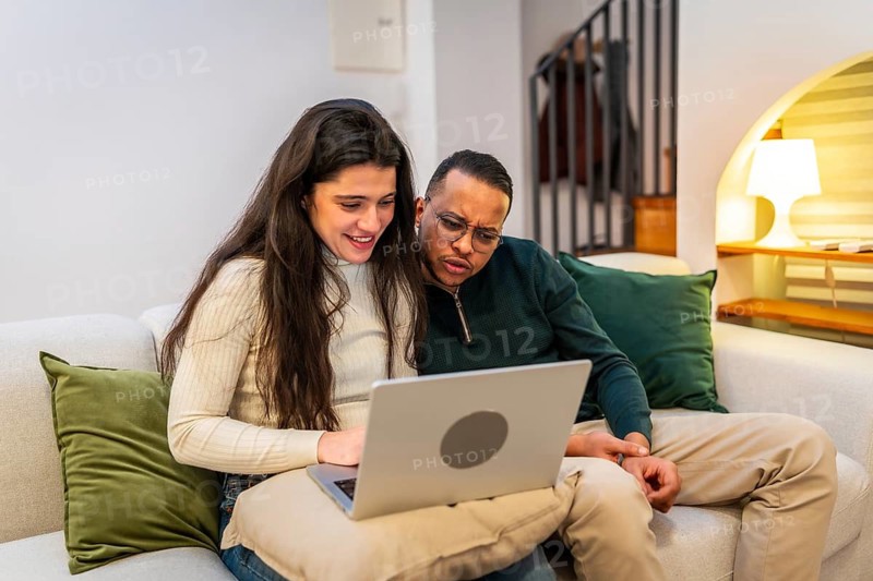 Happy young multiracial couple sitting on a cozy sofa at home, browsing content on a laptop screen, enjoying leisure time and online activities together