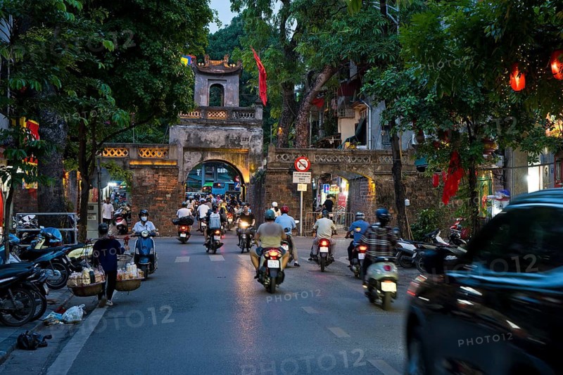 City gate during the evening rush hour on a busy street with mopeds, scooters, motorbikes and passers-by, surrounded by historic buildings and trees, Historic City Gate in the Old Quarter of Hanoi Vietnam