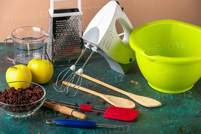 Kitchen utensils, mixer, grater, bowl next to apples and raisins on a table