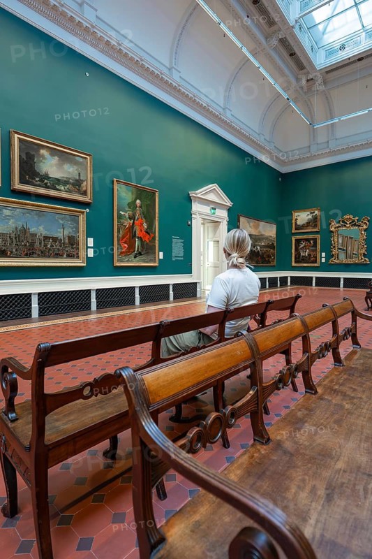 A visitor sits in a gallery in front of paintings surrounded by green walls and wooden benches, National Gallery of Ireland, Dublin, Ireland