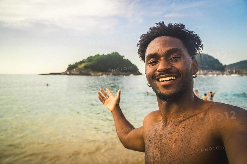 Young black man smiling, taking a selfie on a beach with an island and ocean in the background, enjoying a relaxing summer holiday