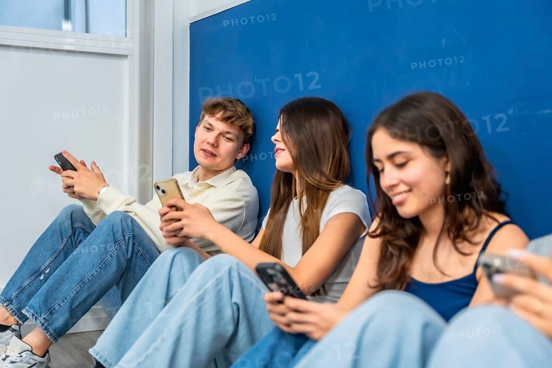 Young students sitting together on the floor against a blue wall, interacting and engaging with their smartphone devices, showcasing modern communication methods and digital reliance