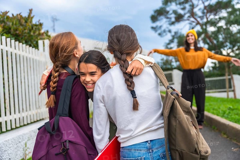 Young girls carrying backpacks and school supplies, smiling and embracing casually on the sidewalk, with a blurred adult woman waiting in the background, signaling a family reunion after school