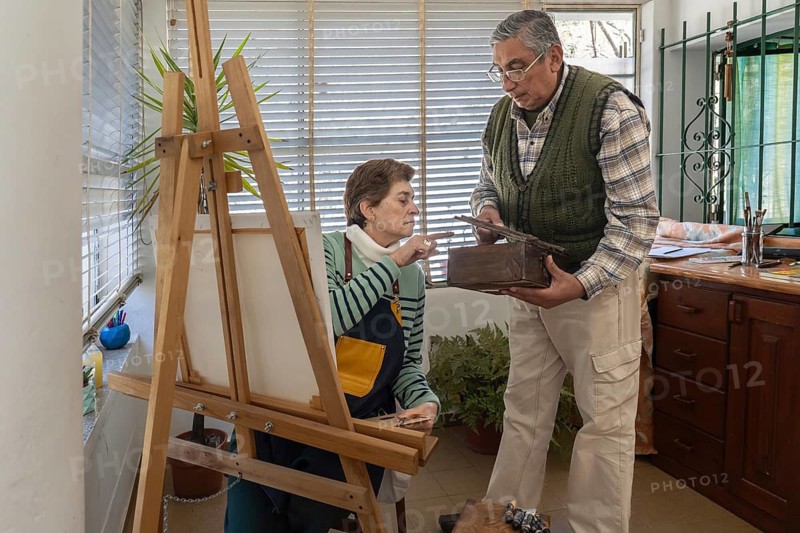 Older woman painter seated at wooden easel examines a reference object held by a man in a home art studio surrounded by plants