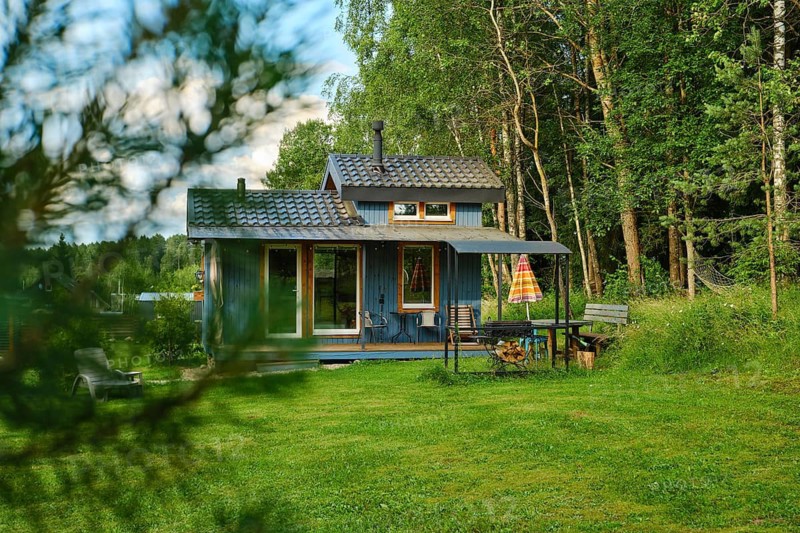 Wooden house stands in green field surrounded by tall trees. Table, grill and chair are set outside. Sunlight shines on the area during daytime
