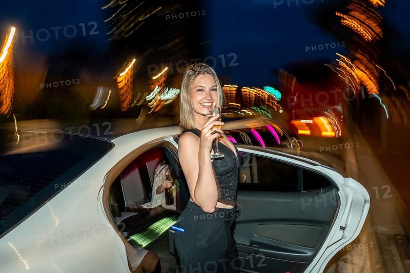 Woman standing beside a white limousine, holding a glass of champagne and smiling, celebrating a special occasion with friends during a vibrant city nightlife experience