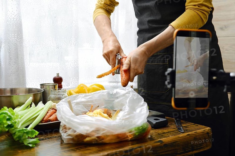 A person is peeling carrots while standing at a wooden table in a kitchen.He records a video on his smartphone for his food blog while explaining the recipe