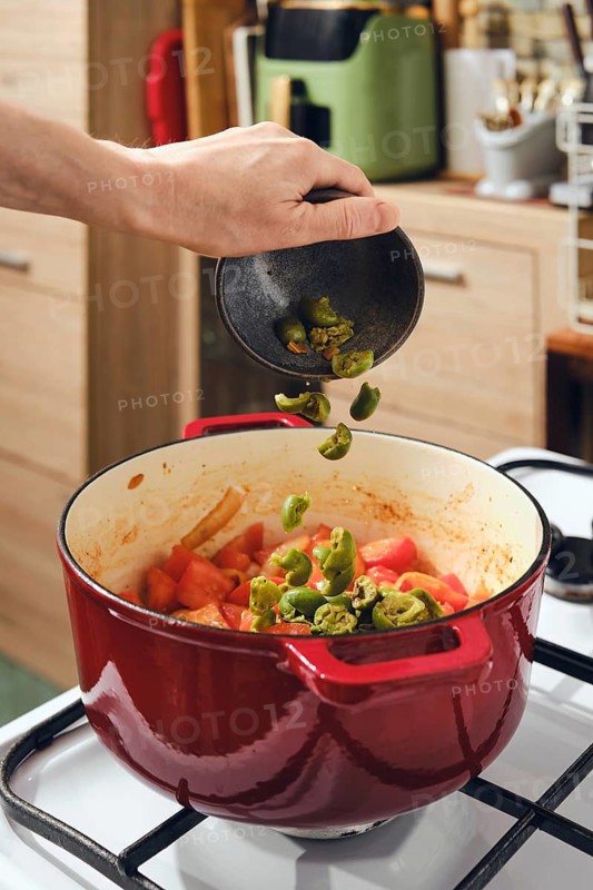 Hand is adding crushed green olives to pot filled with chopped tomatoes and vegetables. Pot sits on stove in cozy kitchen with modern decor and wooden cabinets