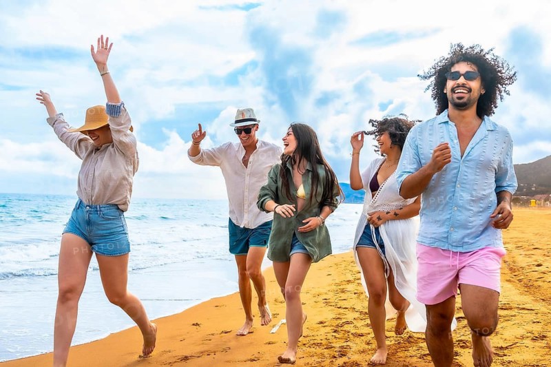 Diverse young friends running barefoot along a sunny sandy beach by the ocean, smiling and laughing together, enjoying carefree summer vacation, freedom and joyful bonding