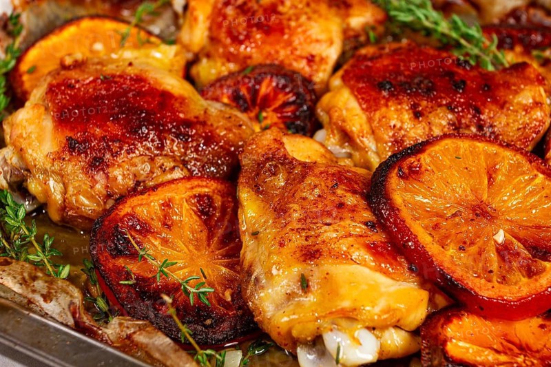 A tray holds cooked chicken thighs topped with orange slices and fresh herbs. The chicken has a rich brown color and is surrounded by juices. This scene shows a kitchen ready for a meal
