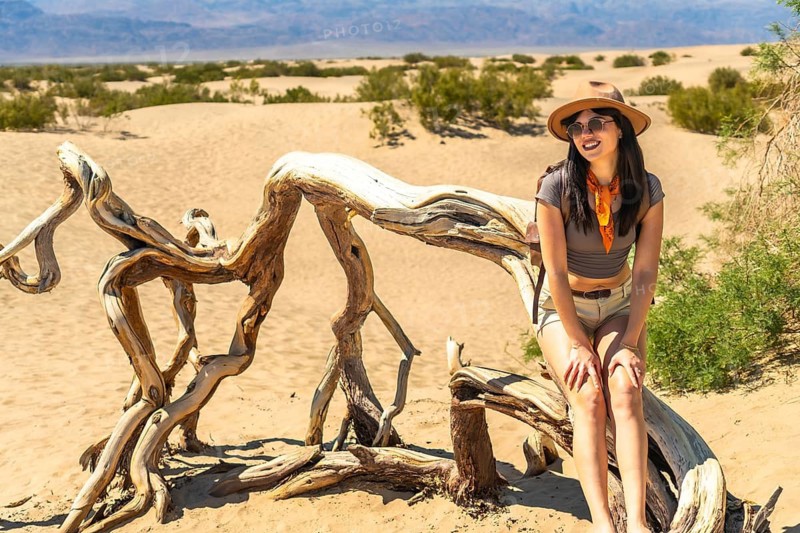 Young woman sitting on a piece of natural driftwood in the sunny mesquite flat sand dunes, smiling and exploring the vast desert landscape during a vibrant travel adventure