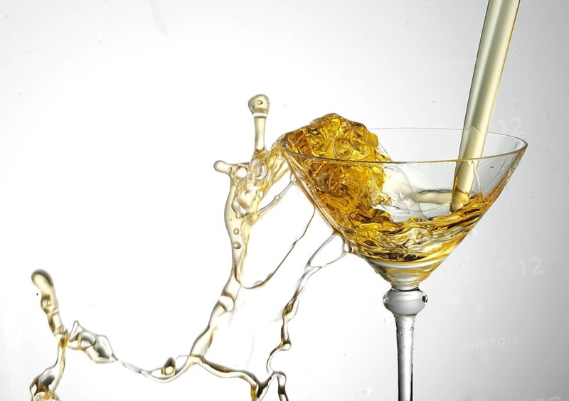 A strong flood of golden yellow liquid pours into a cocktail glass and spills over, studio shot against a white background