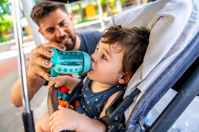 Father holding a drinking bottle for his young child sitting in a stroller, ensuring hydration during an outing and showing nurturing care and bonding
