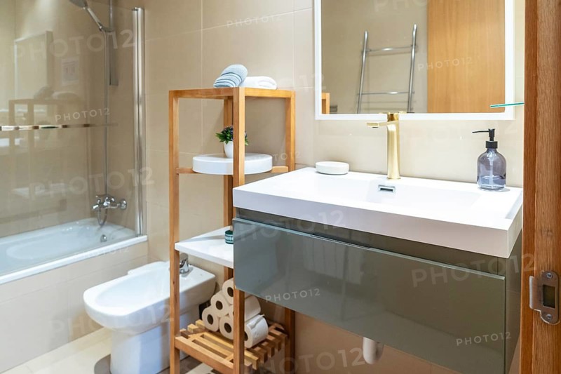 Contemporary bathroom showcasing a white sink with gold faucet, an illuminated mirror, bidet, bathtub with shower, and functional wooden shelving unit