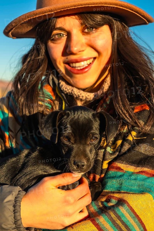 Young woman wearing a hat and a colorful poncho is smiling while holding a small black puppy close, both experiencing a warm sunrise in a desert landscape