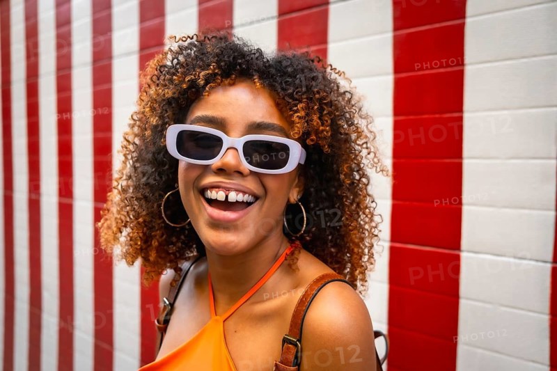 Young woman with curly brown hair wearing purple sunglasses and an orange top, laughing cheerfully against a vibrant red and white striped background, expressing happiness and summer leisure