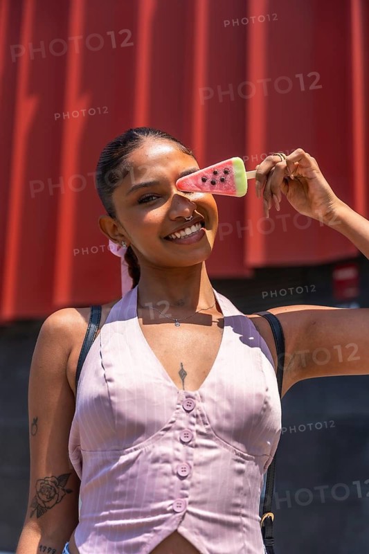 Young woman smiling and holding a watermelon shaped popsicle over her eye, enjoying a refreshing summer treat against a vibrant red background