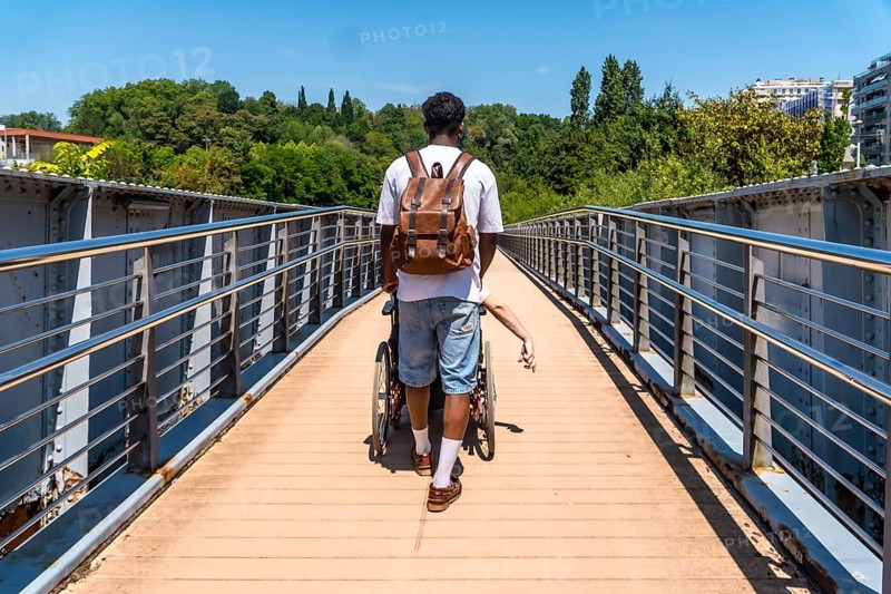 Young man with a backpack pushing a person in a wheelchair across a modern metal bridge on a sunny day, providing support and demonstrating assistance and care for people with mobility challenges